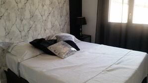 Ein ordentlich bezogenes Bett mit weißer Bettdecke und dekorativen Kissen, eine Wand mit Marmoroptik, ein schwarzer Nachttisch mit Lampe und ein Fenster mit Vorhängen.