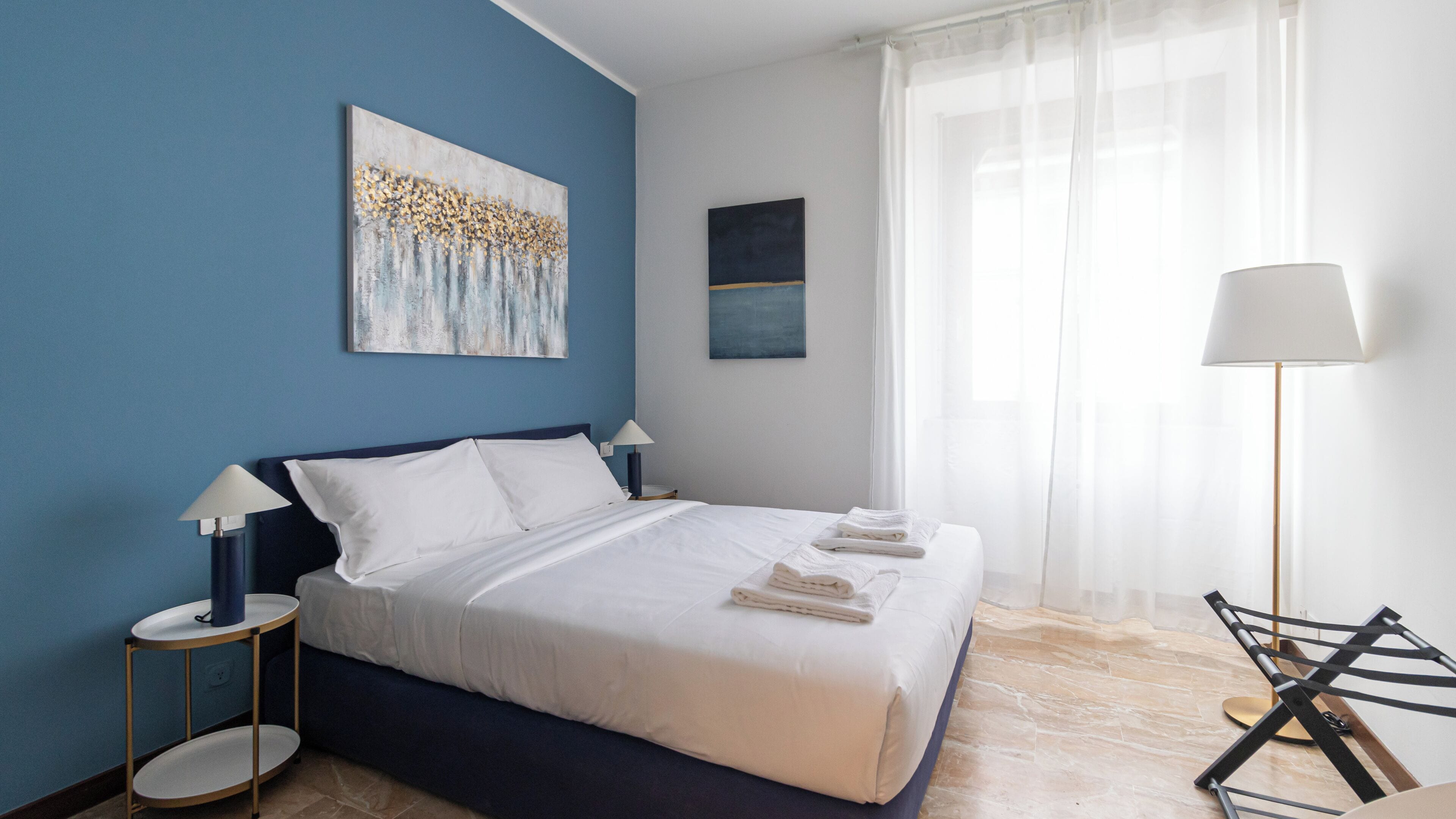 Una habitación de hotel moderna con una cama grande, dos mesitas de noche, una lámpara de pie y obras de arte en las paredes.