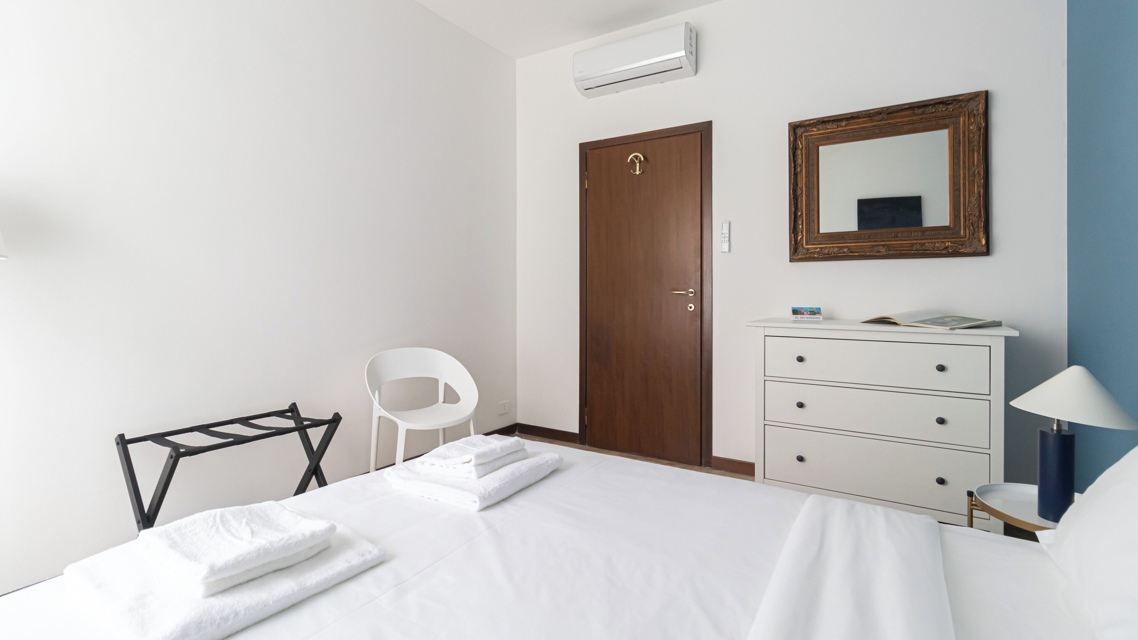 A hotel room with a bed, a wooden door, a white dresser, a mirror, a white chair, and a small table with a lamp.