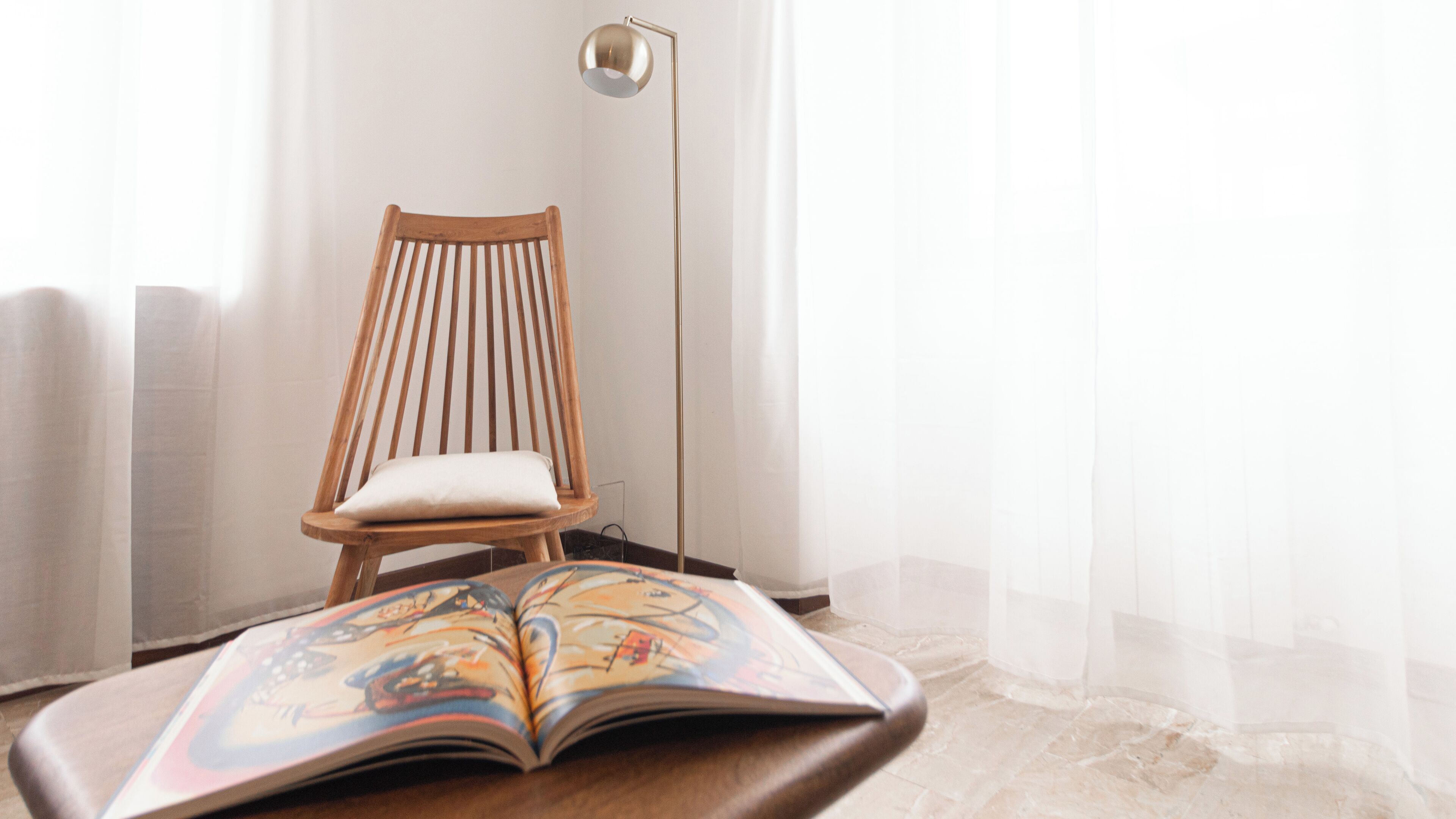Una silla de madera con cojín, un libro abierto con ilustraciones coloridas y una lámpara de pie en una habitación con pared blanca y cortinas translúcidas.