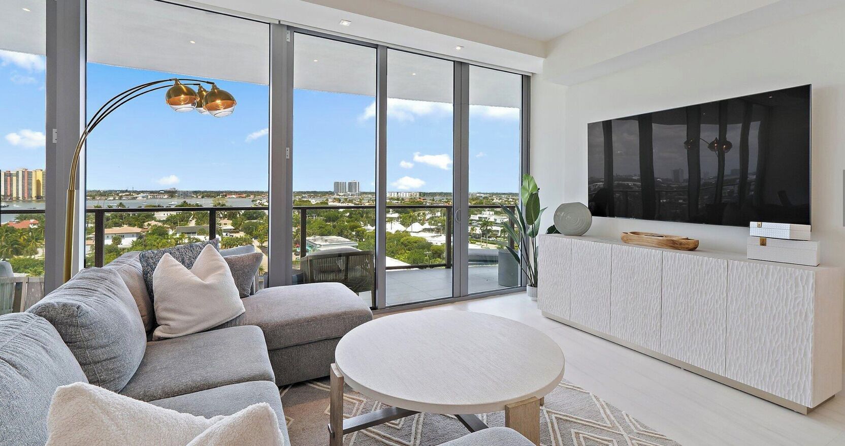 Ein modernes Wohnzimmer mit großem Fernseher, grauem Sofa und Blick auf die Stadt.