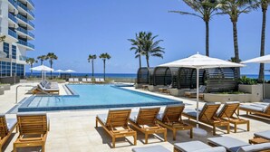 A pool area with lounge chairs, umbrellas, and palm trees. - Luxury Amrit Ocean Resort - 2BDR Condo (Riviera Beach)
