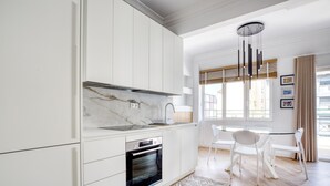 A modern kitchen with white cabinetry, a marble backsplash, and a built-in oven. There is a dining area with white chairs and a table, and a window with blinds. - Whitewood Residence (Athens)