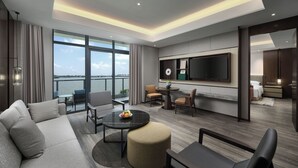 Et moderne hotelværelse med et stort vindue, der giver udsigt over vandet, en sofa, et rundt bord med frugt og et fladskærms-tv monteret på væggen.