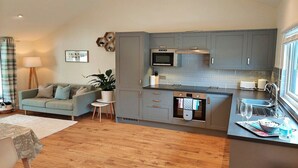 A modern kitchen with grey cabinets, a microwave, oven, and a sink. There is a dining area with a table and chairs, and a sofa. - Cottage in Dartmoor with Private Hot Tub (Bovey Tracey)