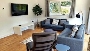 Un salon moderne avec un téléviseur à écran plat, un ensemble de canapés gris, une table basse en bois et une grande plante en pot.