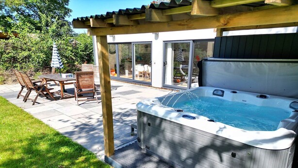 Un bain à remous sous une pergola, une table et des chaises de patio, ainsi qu’une pelouse et une maison en arrière-plan.