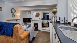 A modern kitchen with white cabinets, a built-in oven, and a sink. There is a wooden shelf with a plant, a mirror, and a clock on the wall. - Cottage in Whitby near Stunning Beaches (Whitby)