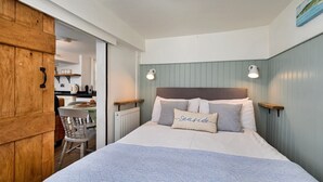 A bedroom with a bed, bedside tables, a wooden door, a chair, and a view of a kitchen area. - Cottage in Whitby near Stunning Beaches (Whitby)