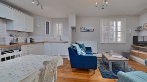Eine moderne Küche mit weißen Schränken, einem Esstisch mit gemustertem Tischlein und einem blauen Sofa. Ein Fernseher steht auf einem Regal.