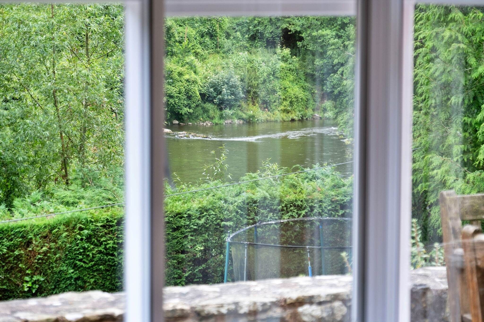 Een rivierlandschap, omgeven door weelderige begroeiing, te zien door een raam met een stenen dakrand en metalen hekwerk.
