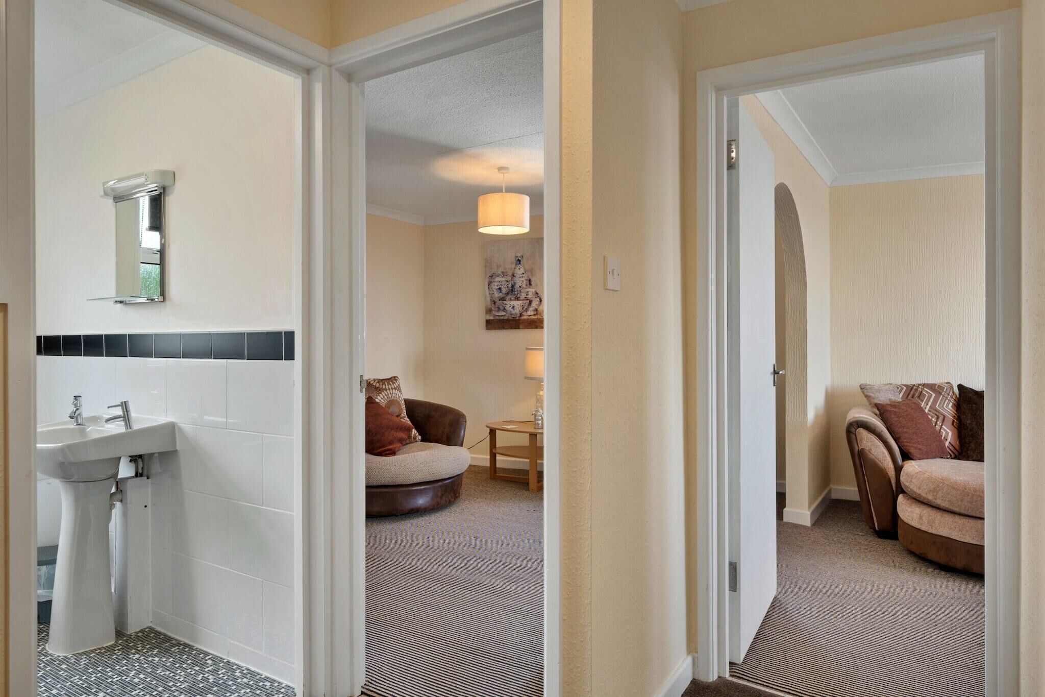 A hotel bathroom with a sink, mirror, and toilet. There is a round chair and a small table with a lamp in the adjacent room.
