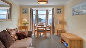 A hotel room with a dining area, a sofa, a small table with a lamp, and a view of the sea.