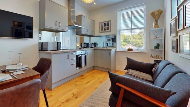 A modern kitchen with grey cabinets, a built-in oven, and a microwave. There is a dining area with a table set for two, and a grey sofa in the living area. - Apartment in Matlock near River Derwent (Matlock)