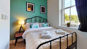 A neatly made bed with a quilt, pillows, and folded towels. There is a bedside lamp, a small table, and a window with curtains. - Apartment in Matlock near River Derwent (Matlock)