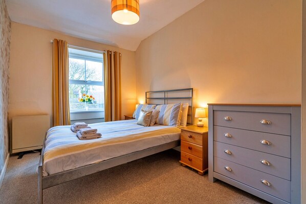 A neatly made bed with pillows and a blanket, a bedside table with a lamp, a dresser with drawers, and a window with curtains.