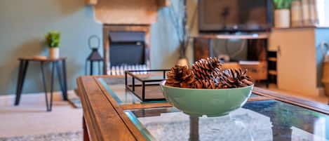 A cozy living room with a glass coffee table, a green bowl with pinecones, a fireplace, and a television.