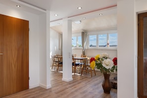 Um quarto espaçoso com porta de madeira, área de jantar com cadeiras e mesa de madeira, e uma janela grande com cortinas.