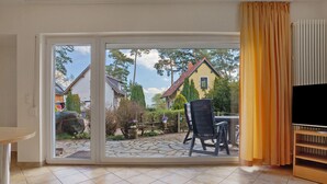 Ein Zimmer mit einer Schiebeglastür, die auf eine Terrasse mit Tisch und Stühlen führt, sowie ein Fernseher und ein Holzschrank.