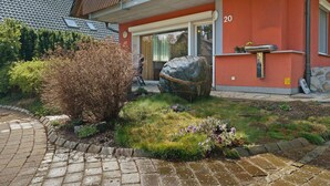 Ein modernes Haus mit roter Fassade, einem Steinweg und einem gepflegten Garten mit verschiedenen Pflanzen und einem großen Felsen.