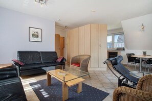 Een moderne woonkamer met een salontafel met glazen blad, een zwarte leren bank, een rieten fauteuil en een eethoek met een ronde tafel.