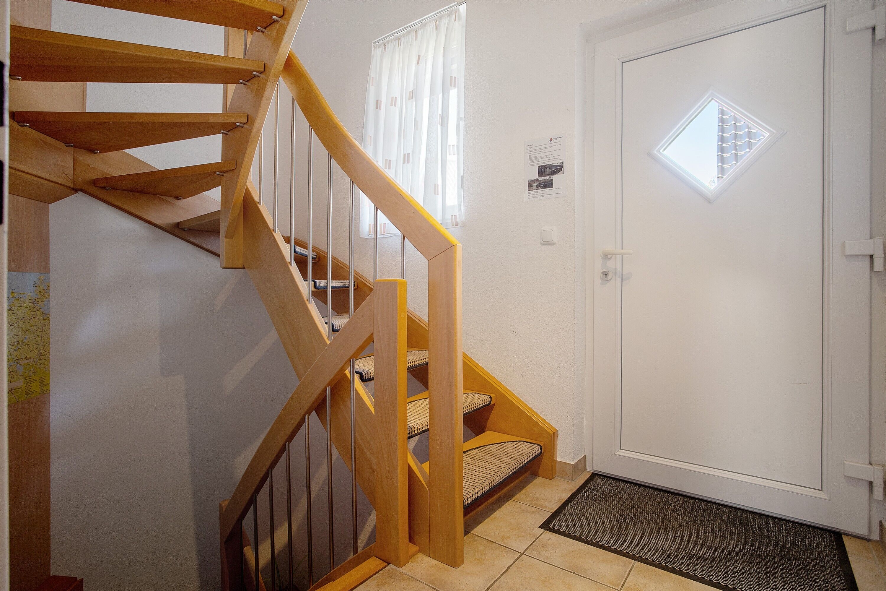 Una escalera de madera que lleva a una plataforma, una puerta blanca con ventana, y una pequeña alfombra en el suelo.