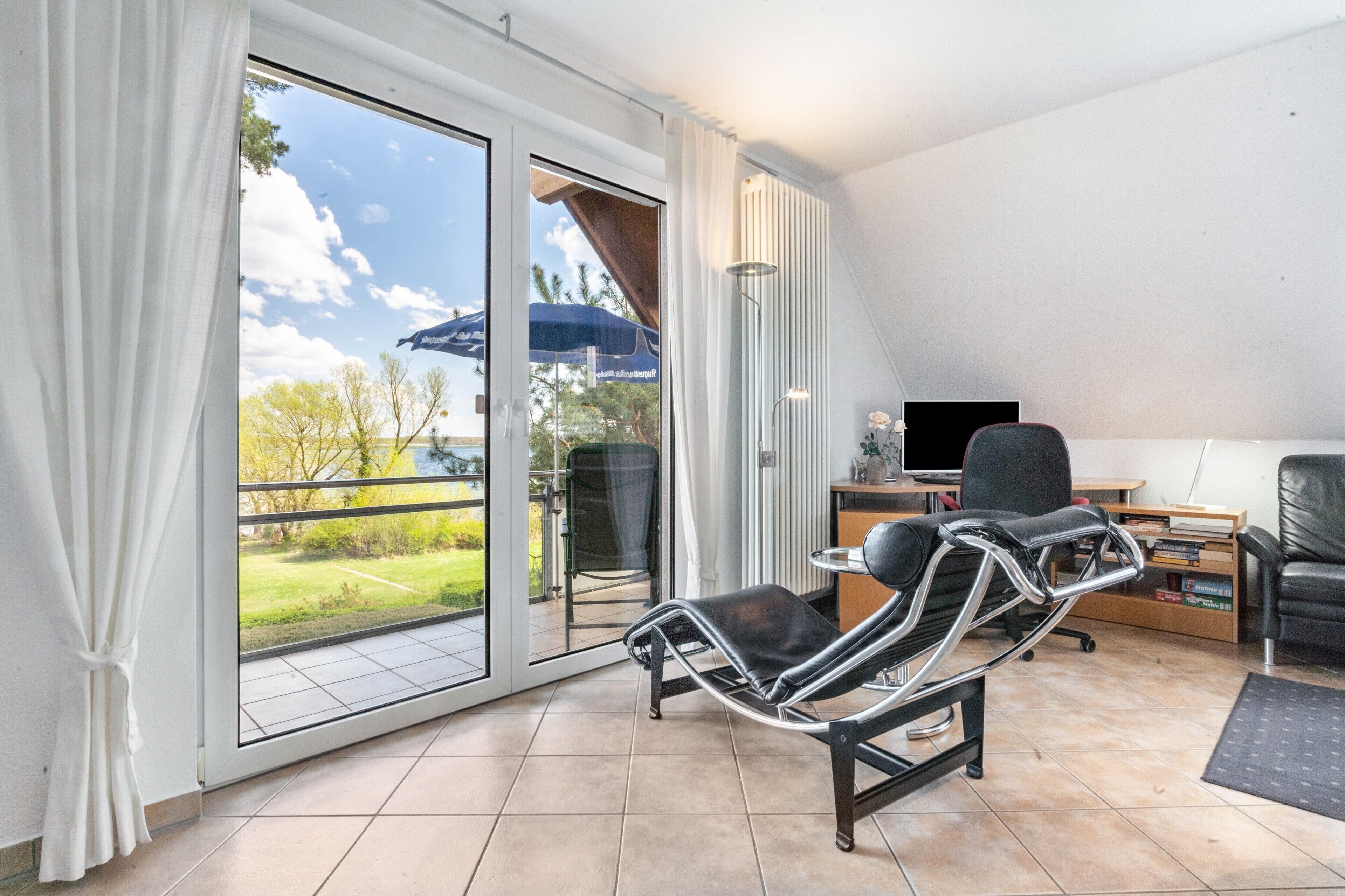 Une pièce avec une porte-fenêtre coulissante donnant sur un balcon, un fauteuil inclinable, un bureau avec un ordinateur et une bibliothèque.