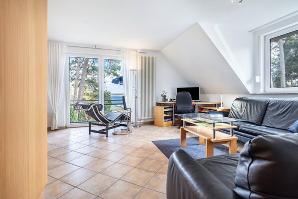 A modern living room with a black leather sofa, a wooden coffee table, a desk with a computer, and a chair. There is a large window with a view of trees and a balcony. - Apartment 'Hs13og mit Müritzblick' with Lake View, Private Garden and Wi-Fi (Röbel/Müritz)
