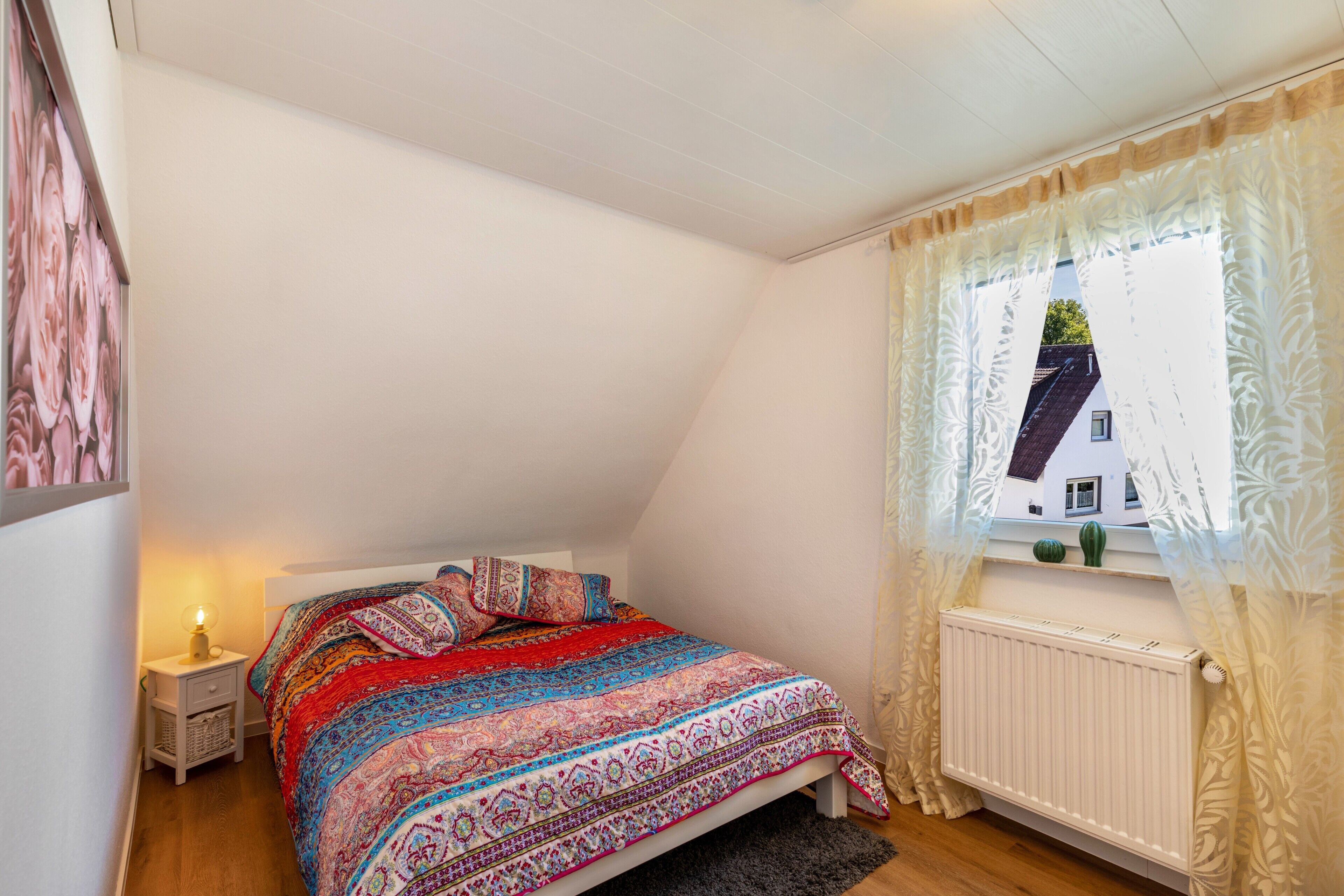 A bedroom with a bed, a nightstand, a window with curtains, and a radiator.