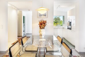 A dining area with a glass table, wicker chairs, a vase with dried flowers, and a pendant light.