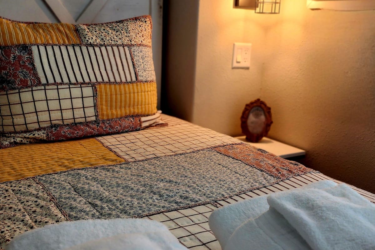 Una cama bien tendida con ropa de cama estampada, una lámpara de noche y una mesita de noche con un reloj.