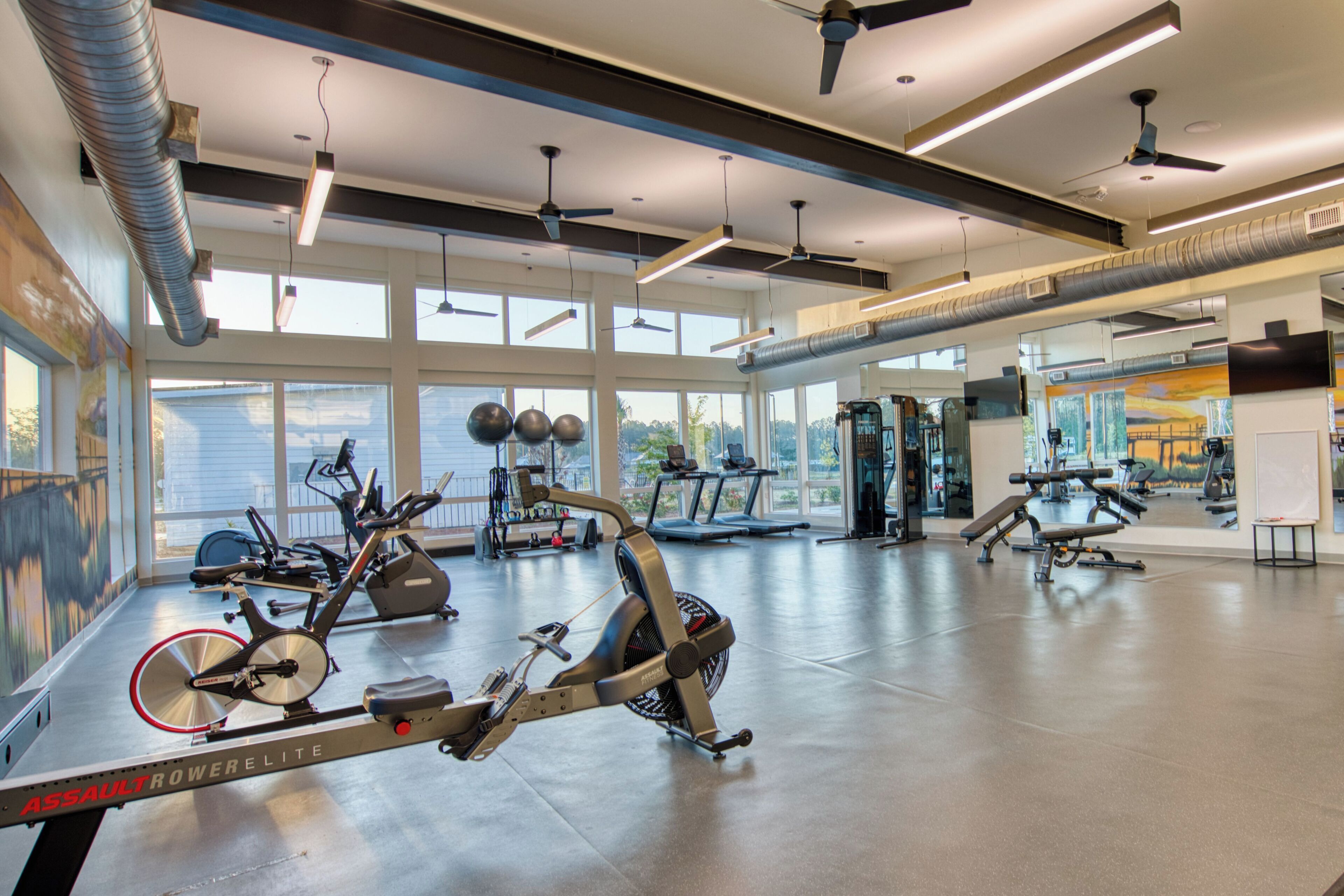 Un gimnasio con equipo de ejercicio, ventanas amplias y ventiladores de techo.