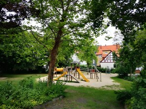 A playground with slides and climbing structures, surrounded by trees and a building with a red roof in the background.