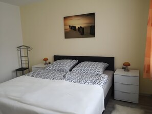 A bedroom with a bed, two bedside tables, a nightstand, a wall-mounted picture, and a hanging coat rack.