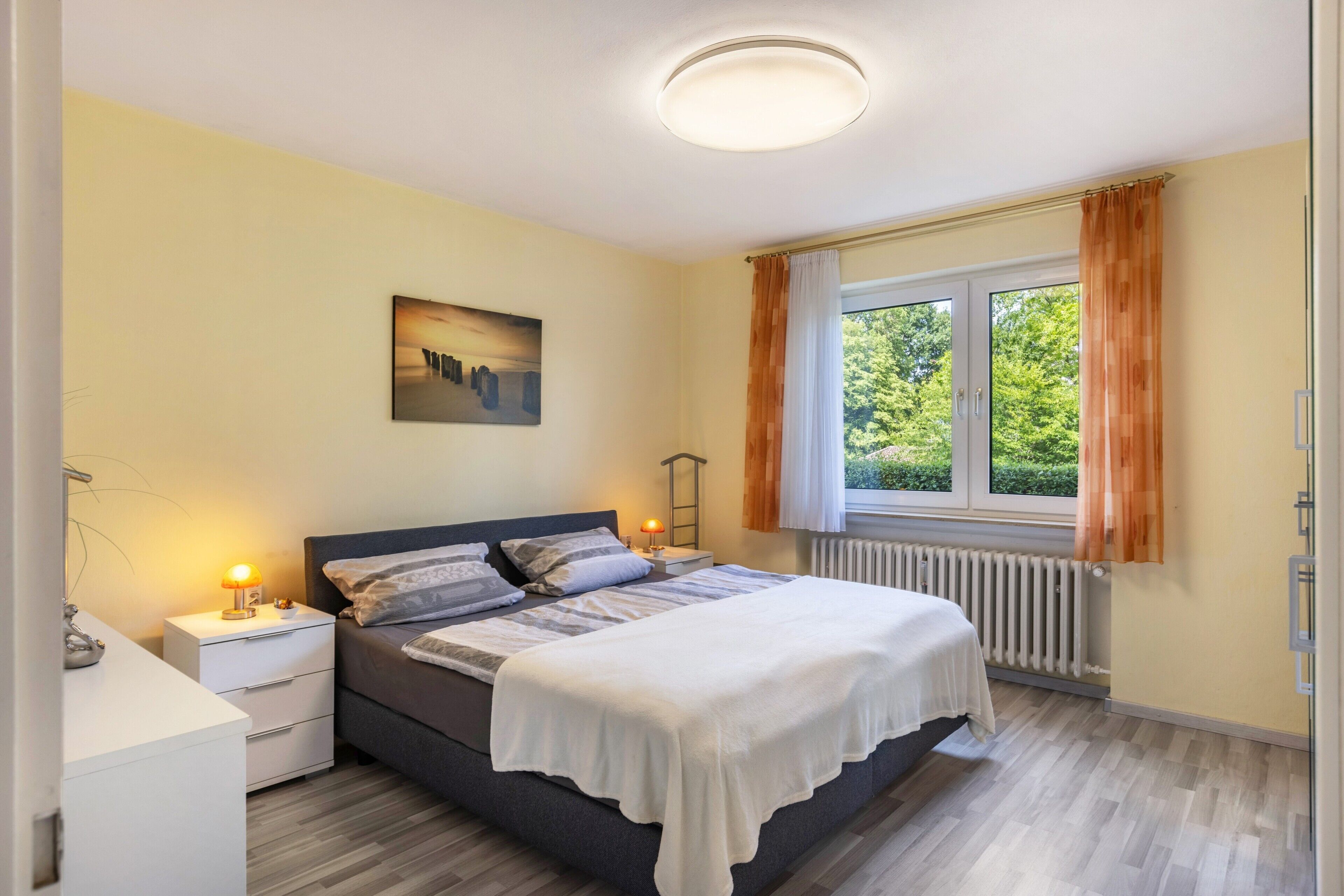 Ein Schlafzimmer mit einem Bett, Nachttischen, einem Fenster mit Vorhängen, einem Heizkörper und einem Bild an der Wand.