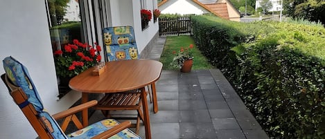A patio with a wooden table and chairs, a small garden, and a view of a white house with a red roof.