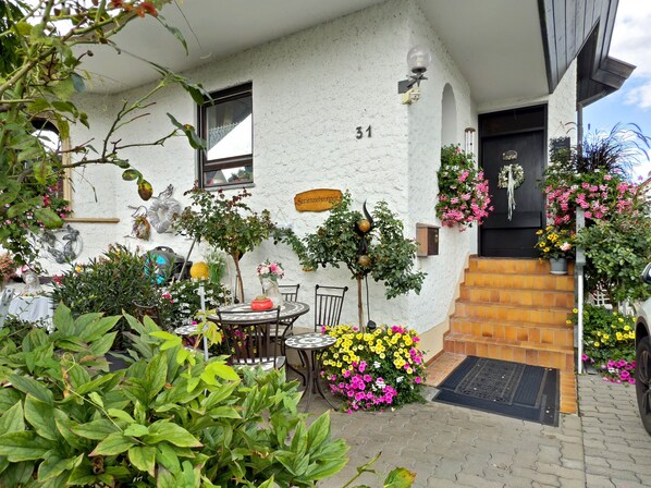 Un edificio blanco con el número 31, un pequeño espacio al aire libre con una mesa y sillas, y diversas macetas con plantas y flores.