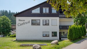 A two-story building with the name 'Haus Gertrud' on the side, a balcony, and a well-maintained lawn with rocks and a tree. - Apartment 'Gertrud' with Shared Terrace and Shared Garden (Schönwald im Schwarzwald)