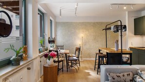 Een moderne woonkamer met een eethoek, een bank met gekleurde kussens, en een keuken met een hangende rek.