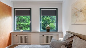 Een slaapkamer met een bed, twee ramen met jaloezieën, een radiator en een schilderij aan de muur.