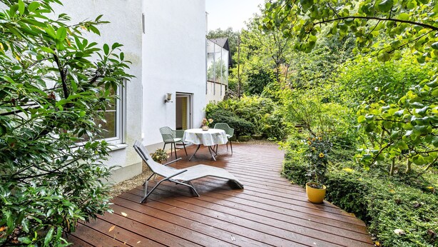A wooden deck with two chairs and a table, surrounded by lush greenery and a white building. - Studio 'Remke' with Private Terrace and Wi-Fi (Starnberg)