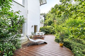 En träveranda med två stolar och ett bord, omgiven av frodig grönska och en vit byggnad.
