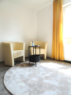 Una stanza con un tappeto rotondo bianco, due poltrone beige, un piccolo tavolino con una statua e una tenda gialla.