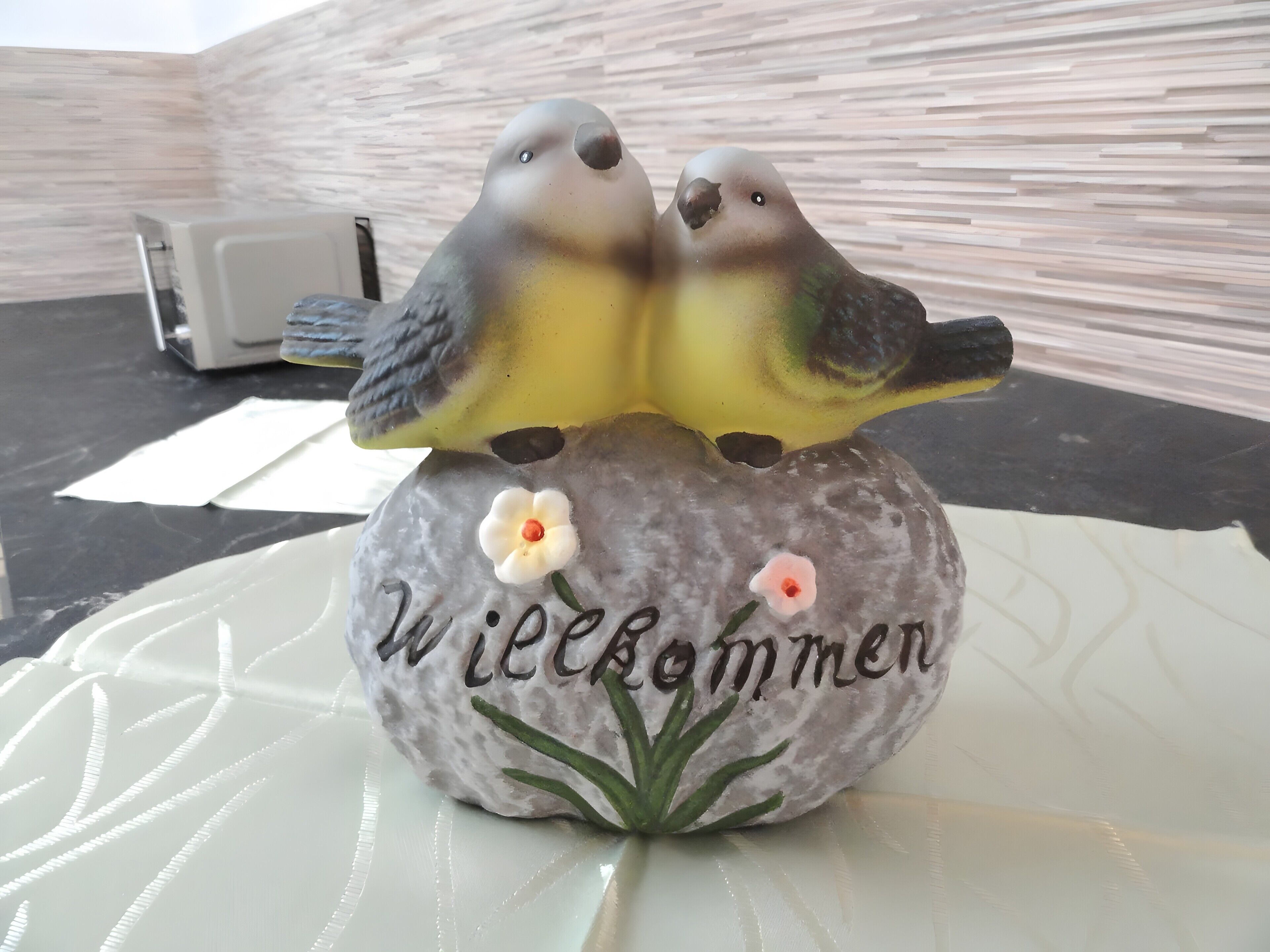 Een decoratief rotspunt met twee vogels en bloemen, en de tekst 'Willkommen'.