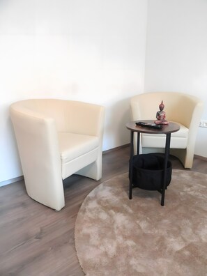 A room with two beige chairs, a small round table with a Buddha statue, and a black bag on the floor.