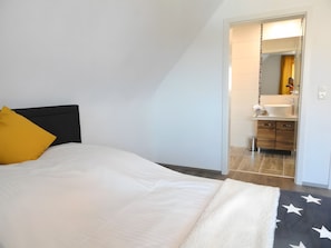 A bedroom with a bed, a bedside table, a mirror, and a bathroom visible through an open door.