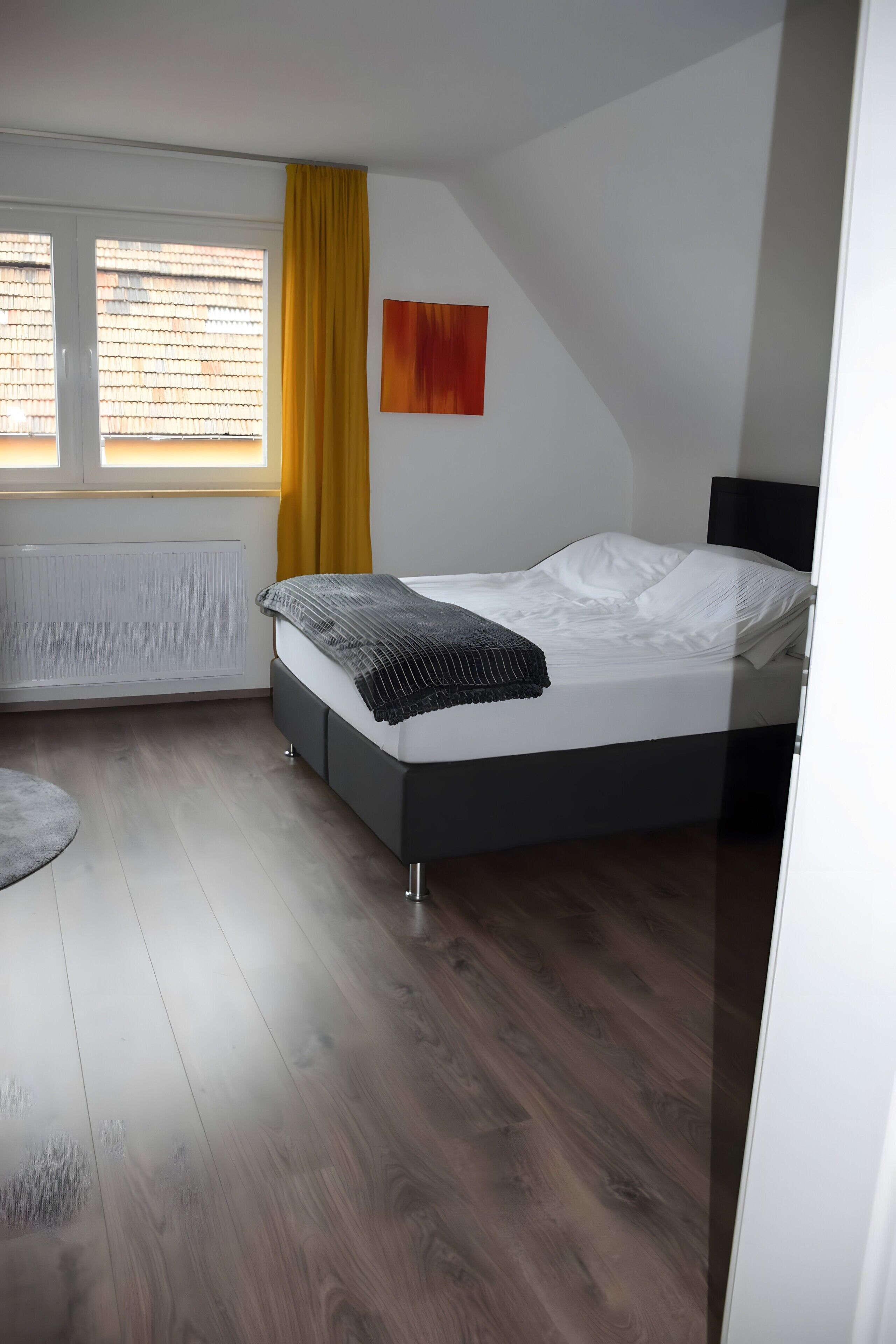 Et soveværelse med en seng, en radiator, et vindue med gardiner og et billede på væggen.
