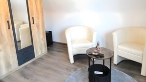 Una habitación con un armario de madera, dos sillones blancos, una mesita redonda y una papelera negra.