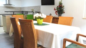 Un comedor con sillas de mimbre, una mesa con mantel blanco, un jarrón verde con flores y un televisor de pantalla plana en la pared.