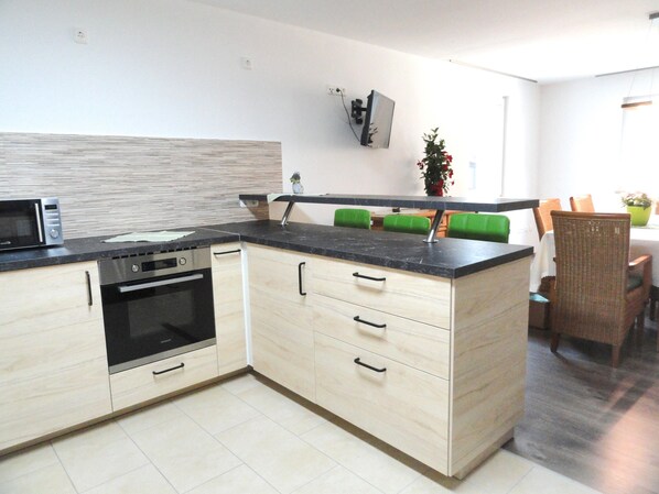 A modern kitchen with built-in appliances, a dining area with green chairs, and a flat-screen TV mounted on the wall.